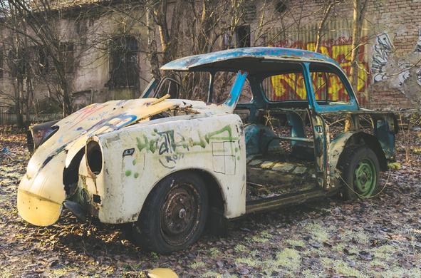A heavily rusted, abandoned car sits among dried leaves next to a graffiti-covered, dilapidated brick building.