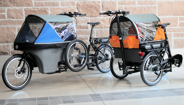 On the left side stands an unfolded, long-john style Wike Electric Salamander cargo bicycle with blue canopy and a "spare wheel attached to the left side of its cargo bay. Front quarter view.

On the right side stands a Wike Electric Salamander cargo bicycle with orange canopy folded into a three-wheeled stroller with the rear wheel, saddle and chainstay now residing on the right side of its cargo bay. Rear quarter view showing the handlebar at the end of the contraption.