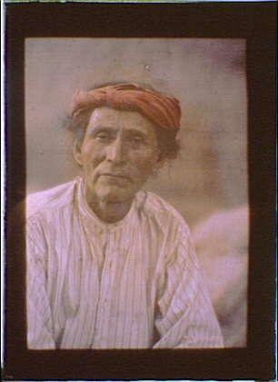 The image depicts a middle-aged man with dark eyes and brown hair, wearing a red turban on his head. He is sitting down and appears to be in deep thought or contemplation. His clothing consists of a striped shirt that looks like it's made from coarse fabric such as cotton. The background behind him features soft-focused natural scenery which could possibly indicate an outdoor setting. It has a sepia tone, suggesting the photograph might have been taken during the late 19th century when autochrome photography was popular for its vibrant colors and ability to create realistic images.