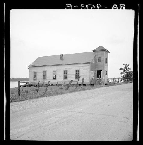 The image depicts a black and white photograph of an old wooden building with architectural features reminiscent of rural American vernacular architecture from the late 19th or early 20th century. The structure has a simple rectangular form, two stories in height, and appears to be weathered by time, suggesting it may have been used for various purposes over its history.

The main body of the building is topped with what seems like a gabled roof, while an additional section on one end rises higher, possibly functioning as a tower or bell-cord. This upper portion has vertical siding and features such as louvered windows which are common in schoolhouses or community buildings from that era.

Surrounding the structure is a rustic setting with a dirt road leading up to it and extending away into the distance on the right side of the frame, suggesting remote accessibility. To the left, there's an open field with sparse vegetation, indicative of rural America during this period. A wooden fence or barrier runs parallel along part of the building’s perimeter.

In the background, a body of water is visible to the far left, hinting at proximity to a river like the Mississippi mentioned in the additional information provided for context. The overall condition and style of the photograph suggest it might be from an archive collection documenting rural American life or architectural history during that time period.