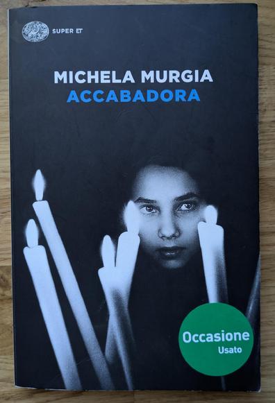 La copertina del libro Accabadora di Michela Murgia presenta un'atmosfera cupa e suggestiva, dominata dai colori nero e bianco.
Lo sfondo è interamente nero. In alto a sinistra si trova il logo dell'editore "SUPER ET". Il nome dell'autrice, MICHELA MURGIA, è scritto in bianco in alto, mentre il titolo del romanzo, ACCABADORA, è in un blu acceso, posizionato centralmente.
Nella metà inferiore della copertina, l'immagine è divisa tra un gruppo di candele bianche accese a sinistra e il volto di una giovane persona (probabilmente una ragazza) a destra, che guarda direttamente l'osservatore con un'espressione intensa e gli occhi spalancati. Le fiamme delle candele e il volto sono illuminati, risaltando sul fondo scuro
