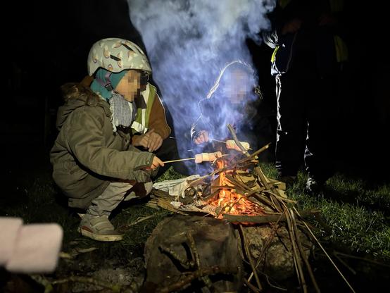 Photo de nuit. Deux enfants au bord d'un foyer de feu de bois naissant. L'enfant à gauche porte un casque de vélo, l'autre est masqué par la fumée. Les deux tiennent une brochette de guimauves au dessus du feu. On distingue deux adultes en arrière plan.