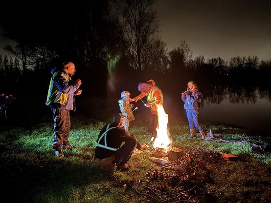 Photo de nuit. Au bord d'un étang noir dans lequel se reflètent les arbres de la ripisylve,  cinq personnes sont autour d'un foyer de feu dans l'herbe. Le feu de petit bois a de belles flammes. Les personnes autour du feu tiennent des brochettes de guimauve et portent des tenues cyclistes (gilet réfléchissants, et un enfant est casqué).