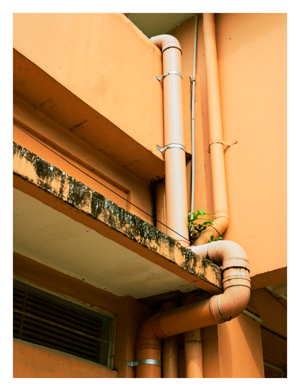 An orange building exterior featuring large, matching drainage pipes running vertically and horizontally along the walls. A weathered, dark-stained concrete ledge provides a textured contrast to the smooth painted surfaces, and a small green plant can be seen growing out of a crevice near the pipe connections. - Google Gemini 3 Pro Preview
