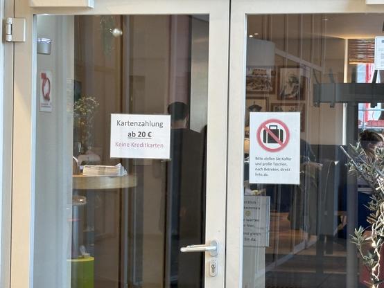 The image shows a glass door with two prominent signs. One sign states that card payments are accepted starting from 20 euros and that credit cards are not accepted. The second sign prohibits bringing large bags and luggage inside, instructing visitors to leave them to