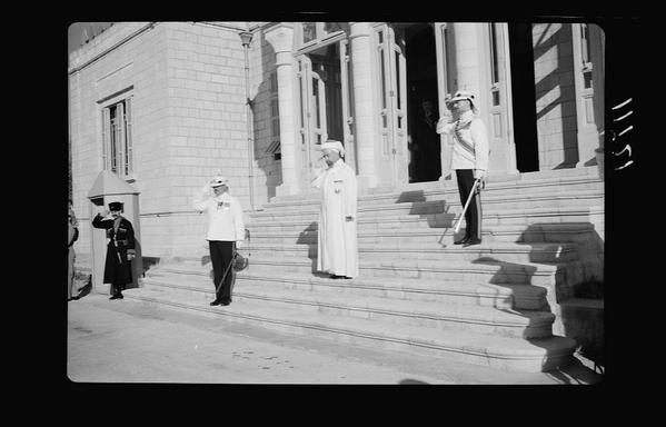 The black and white photograph depicts a formal occasion taking place outside of what appears to be an official government building. There are four individuals in the frame, all dressed in ceremonial uniforms with hats or caps. On the left side, one person is standing guard-like near the entrance steps, wearing dark robes and holding a stick that could be used for directing traffic. In the center-left area, two figures stand on the stairs; both wear white traditional Arab attire complemented by tall cylindrical hats known as keffiyehs or agal caps with a central knot called ghutrah. They are saluting in unison, suggesting respect towards someone off-camera or perhaps during a national anthem moment.

To their right stands another figure also dressed formally but in lighter-toned clothing and holding what appears to be an ornate staff which is often associated with ceremonial duties like warding off evil spirits or as part of traditional guards. The fourth person on the far right side, only partially visible at this angle, seems similarly attired.

The architecture includes steps leading up to a building entrance flanked by columns and windows that reflect sunlight due to its bright surroundings indicative of daytime conditions. This could be during an event commemorating historical or national significance given their formal attire and gestures of salute which suggests respect for someone important being present off-came [...]