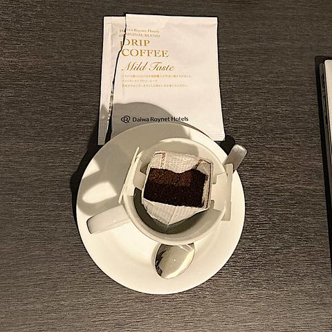 a small cup and saucer with a packet of coffee grounds suspended in the cup by paper “wings” hanging from the edges of the cup. a torn package labeled Drip Coffee Mild Taste lies above the cup.