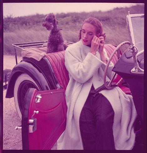 In this color photograph, a woman is seated in the passenger seat of an old-fashioned convertible automobile with red leather interior. She's dressed elegantly in a white coat and black pants, accessorized with pearl earrings and holding a silver cigarette case or small clutch bag. Her hair is styled in soft waves typical of mid-20th-century fashion trends. The woman appears to be applying lipstick while looking down at her hand, which holds the cigarette holder.

A dark-colored poodle wearing a collar sits on an elevated platform behind her headrest, gazing into the distance with its tongue out slightly. Next to them is a large red suitcase that rests on top of another seat in front of the car door handle and lock mechanism.

The background features blurred natural scenery suggesting they're parked outdoors near some bushes or grassy terrain, giving an impression of leisurely travel during vacation times. The overall atmosphere exudes sophistication and grace typical of fashion photography from around 1950-60s America.