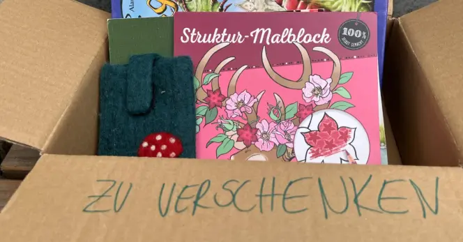 Ein geöffneter kleiner Karton. Auf einer Lasche steht „ZU VERSCHENKEN“. Im Karton stehen Kinder- und Malbücher und eine Handyhülle aus Filz.