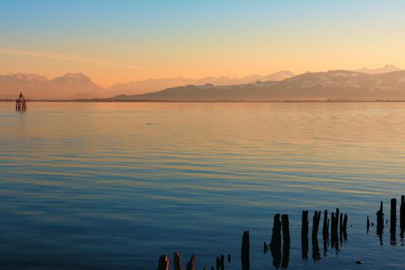 view over a large lake with mountains on the far shore. a row of wooden poles in the water on the lower right. it's evening, the water close to us is dark blue, in the distance it is orange and so are the mountains. the sky above the mountains is light blue. somewhat kitschy.