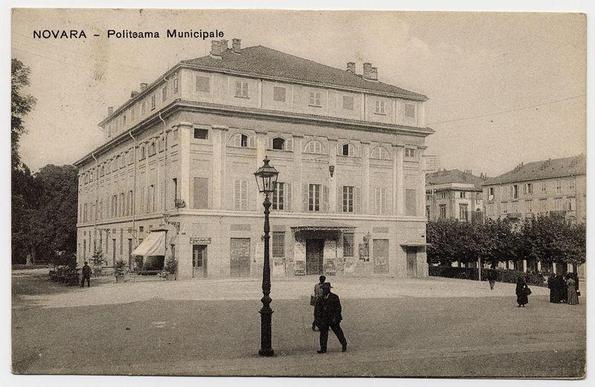 The photo depicts an old, stately municipal building in Novara. The architecture of the building is grand and classical with a symmetrical facade featuring multiple windows framed by white shutters, and centered on columns that support an upper balcony-like structure. There are two chimneys visible at the top, suggesting it might have been used for heating during colder months.

In front of the building stands a vintage street lamp post, indicative of historical urban planning or maintenance practices before modern lighting technology became prevalent. The ground appears to be paved with cobblestones which were common in older city areas.

There are several people scattered around the scene; one person is walking on the left side near what seems like an entrance that may have been a café based on signage, and others appear engaged in conversation or casual strolls along the street. The clothing style of the individuals suggests this photo could be from late 19th to early 20th century.

In front of the building is an open space with benches aligned against what appears to be another establishment with a sign reading 'Caffè & Osteria'. This indicates that in addition to serving as municipal offices, it may also have served social or community functions. The environment gives off an air of tranquility and order.

The photo has some text on the top left corner stating "NOVARA - Politeama Municipale", which translates from It [...]