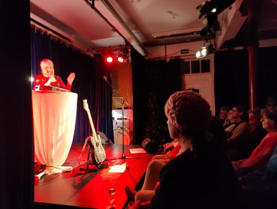 Ich, hinter einem Stehtisch auf der Bühne mit meiner Hirnmütze und im Vordergrund ein Mann im Publikum mit meinem " Leihirn" auf dem Kopf.