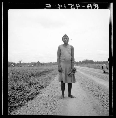 The black and white photograph depicts a woman standing on a dirt road, with rural farmhouses and cultivated fields visible in the background. The subject is barefooted, wearing an apron-like dress that reaches mid-calf length, holding what appears to be food items or possibly religious symbols, such as candles or small statues. Her expression is neutral but contemplative.
The image captures a moment of everyday life within rural America during a period when people were likely engaged in manual labor and agricultural work. The overall mood conveyed by the photograph suggests hardship, resilience, and simplicity associated with this era's lifestyle.