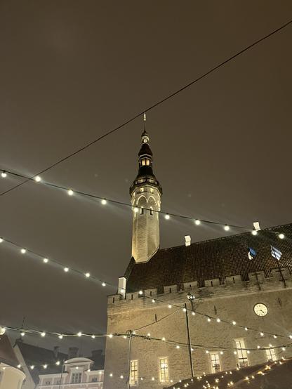 A tall tower with a pointed spire is illuminated against a dark sky. The building features a stone facade and several lit windows. String lights are strung in the foreground, adding a festive atmosphere.