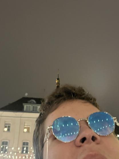 A close-up selfie showing part of a person's face wearing round, reflective blue glasses. The background is blurred with building outlines and festive lights.