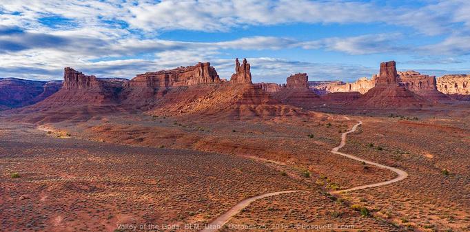 An aerial view of the edge of southwest US desert canyon country with steep redrock cliffs, buttes, and mesas with debris below at the angle of repose. In the foreground a rolling landscape of red dirt with small green shrubs. A narrow dirt road roams from bottom center to middle right. A pronounced wash "flows" from middle left to lower right. Blue sky with both white and dark clouds.
©BosqueBill.com