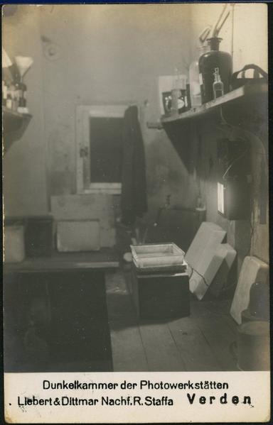 A black and white postcard showing the inside of a photographer's developing chamber. There is a curtained window, a desk, various bottles on a shelf, various pans for holding liquids and chemicals.