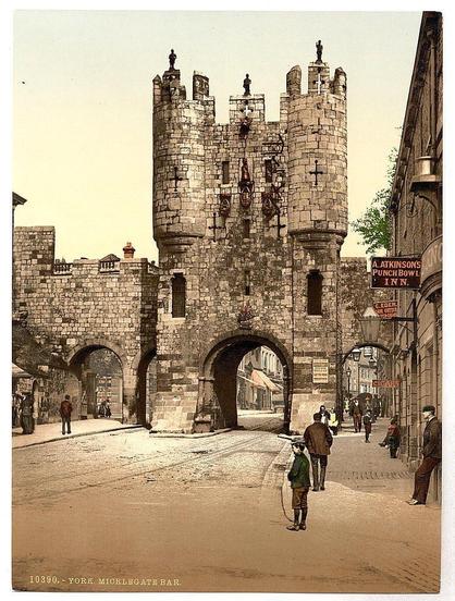 The image depicts a historic stone archway known as Micklegate Bar in the city of York, England. The structure has four robust towers with crenellations and features Gothic architectural elements such as pointed arches. Several people are scattered around the area; some appear to be walking while others stand still or interact with each other. On one side of the bar is a sign that reads "A. Atkinson's Punch Bowl I.N.N." indicating an establishment for travelers during their stay in York. The overall atmosphere suggests this was taken between approximately 1890 and 1900, capturing daily life at Micklegate Bar with vintage attire worn by individuals indicative of the era.