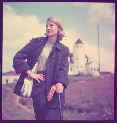 A woman stands on a grassy field with her hand resting against what appears to be the handle of an umbrella. She is dressed in period-appropriate clothing, featuring a grey coat over a light-colored dress or blouse and dark pants. The outfit includes gloves that are white and appear to have fur trim at the wrists. Her hair is styled into waves typical for the early 20th century.

Behind her stands a prominent building with architectural features such as a tower-like structure topped by what seems like cupolas, possibly indicative of an institutional or governmental function. The sky above has patches of clouds but predominantly appears to be clear and bright.