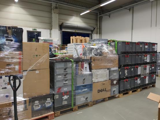 Ein Foto einer mit kaltweißem Licht erleuchteten Lagerhalle. Im Vordergrund sind 5 Reihen mit Europaletten zu sehen. Bepackt mit Kartons, Euroboxen, Waschmaschinen, Druckern usw.
Die Paletten sind einfoliert und mit Spanngurten gesichert. Im Hintergrund ist ein offenes Rolltor und ein Notausgang erkennbar.
