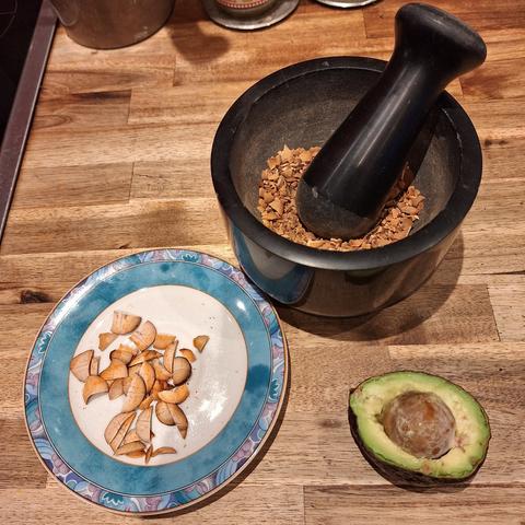 Auf einer braunen Arbeitsplatte aus Holz steht ein schwarzer Mörser mit kleinen braunen Stücken vom Avocado-Kern. Links unterhalb auf einem kleinen weißen Teller mit blauem Rand liegt der Kern in Scheiben geschnitten und rechts davor eine halbe aufgeschnittene gelbgrüne Avocado mit Kern.