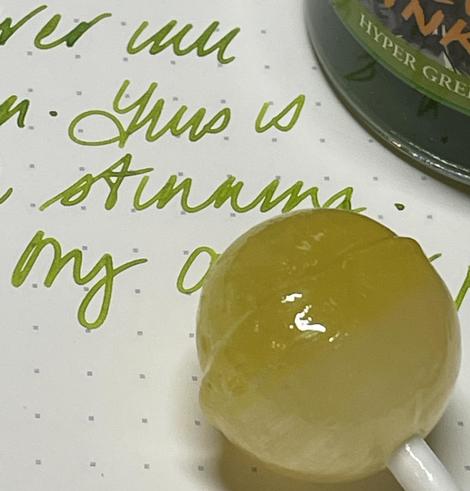 An image featuring a yellow lollipop resting on a dotted notebook. Nearby, there's handwritten cursive text in green ink. A jar labeled "HYPER GREEN" is partially visible in the background.