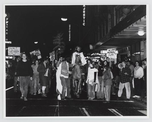 A black and white photograph captures a group of demonstrators on a city street at night, likely during the Vietnam War era given the protest signs. The protesters are holding various placards with messages such as "We want moral films" and political slogans like “Some people’s Vietnam." A central figure is mounted atop a horse amidst other marchers carrying banners that read "Murder! In our name - Vietnam," indicating active participation in anti-war protests of the time period. The image exudes an atmosphere of activism, with protesters showing determination through their body language and expressions as they navigate the urban environment together.