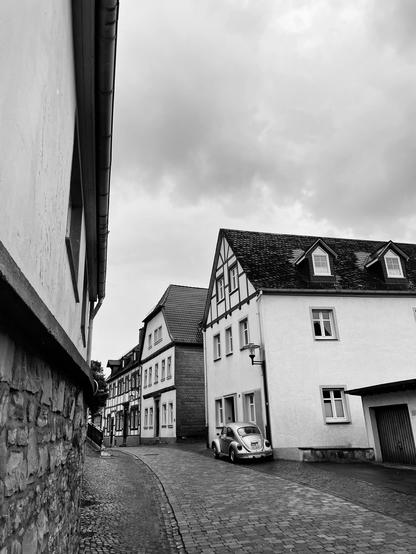 A narrow, cobblestone street in a quaint village featuring a mix of traditional and modern buildings, with overcast skies overhead. A vintage car is parked alongside the houses. The scene is in black and white.