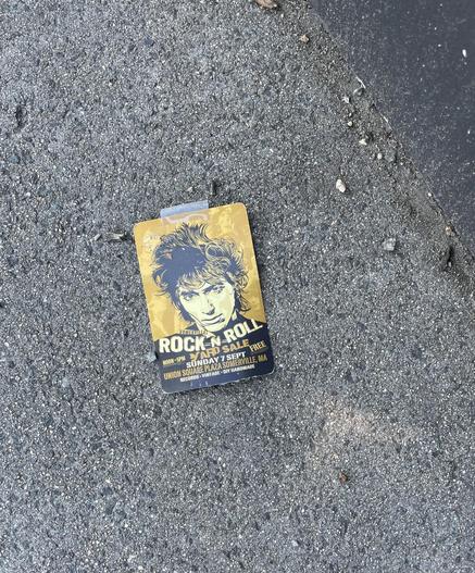 An advertising card lying on gravelly pavement. Upper 2/3 of the card has a drawing of a head shot of a guy with shaggy/spiky hair, eyes glowering up at us from his slightly lowered head, on a mustard colored background bearing faint images of guys of similar ilk singing into microphones. Below pic, printed in off-white, text ras flows:"Somerville" in small text at an angle, as if added as an afterthought, then "ROCK-N-ROLL" on the next line in large text curving under the guy's chin, then below that several lines of smaller text in stencil-like block letters in varying shades of mustard and off-white "Noon to 5PM yard sale FREE, Union Square Plaze Somerville, MA, Records * Vintage * DIY Handmade" The piece of tape that failed to keep it where it was afixed is still attached at top of card.