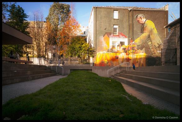 Wide-angle photograph of Bishop Lucey Park in Cork showing a sunlit public space with central grass strip, large-scale mural on a tan-coloured building depicting a man in a yellow shirt working on a table draped in orange cloth with a house illustration below, concrete steps and pathways flanking the grass, a small child running on the right, autumn trees with orange foliage, historic city buildings visible in background under clear blue sky.