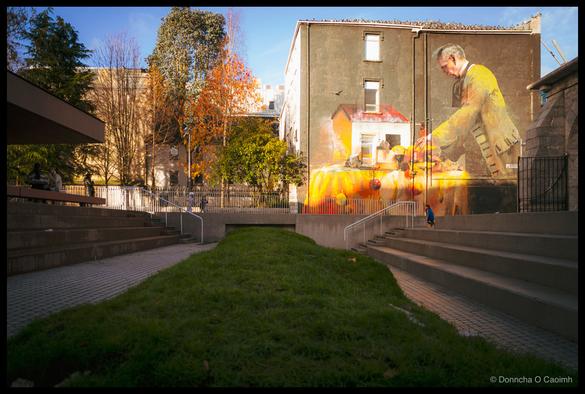 Wide-angle photograph of Bishop Lucey Park in Cork showing a sunlit public space with central grass strip, large-scale mural on a tan-coloured building depicting a man in a yellow shirt working on a table draped in orange cloth with a house illustration below, concrete steps and pathways flanking the grass, a small child running on the right, autumn trees with orange foliage, historic city buildings visible in background under clear blue sky.