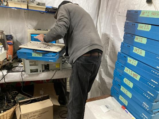 A tall stack of bright blue SGI Indy computers sits along one wall. Opposite of that an elderly man hooks up one of the Indy's to a CRT monitor and keyboard.