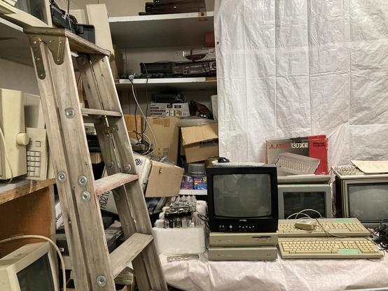 Another corner of the computer lab. A white tarp hangs from the wall, and in front of it are Atari ST computers and monitors and televisions. To the left are dozens of macintosh computers in varying condition. A wooden ladder rests against them.