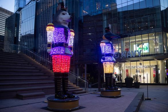 Two large, colorful light displays resembling toy soldiers are positioned outside of an Old Navy store. One features a wolf's head, while the other has a bird's head. The figures are illuminated with vibrant lights, and the setting is urban, with glass buildings
