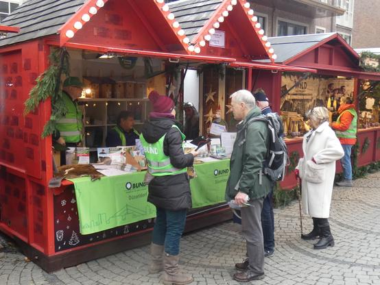 Foto von der mit BUND-Banner geschmückten Weihnachtshütte. Davor steht eine BUND-Aktive mit Weste im Gespräch mit Bürger*innen. (Foto: Klaus Kurtz)