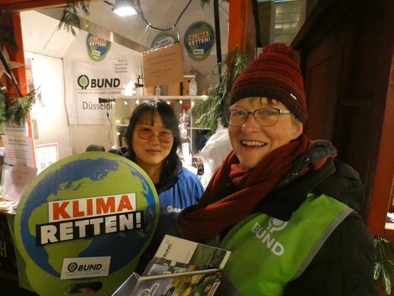 Nahaufnahme von zwei BUND-Aktiven in der Weihnachtshütte. Im Vordergrund zudem ein BUND-Schild in Form der Erde, auf dem "Klima retten!" steht. (Foto: Klaus Kurtz)