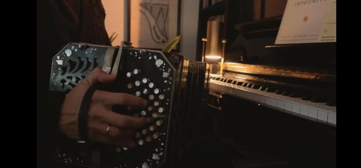 A softly lit scene showing a person playing a bandoneon, we just see the hand, with a dark wood piano and a music score on the right of the frame.