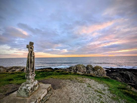 Un calvaire moderne en granit au bord d'une falaise avec vue sur l'océan et le coucher de soleil très obstrué. Les couleurs dans un camaïeu de bleu sont jolies.