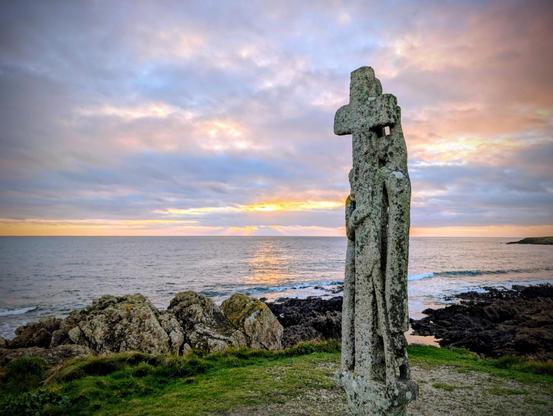 Un calvaire moderne en granit au bord d'une falaise avec vue sur l'océan et le coucher de soleil très obstrué. Les couleurs dans un camaïeu de bleu sont jolies.
