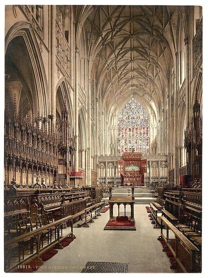 The image depicts the majestic interior of a cathedral with Gothic architectural elements. The viewer's perspective is from within, looking down an expansive nave towards the altar at the far end. On either side are rows upon rows of wooden benches and choir stalls in dark hues, indicating their use by clergy or choirs during services. Above these seating areas runs ornate stained glass windows that fill the space with colorful light when sunlight streams through them.

The high vaulted ceilings add a sense of grandeur to the scene, adorned with intricate tracery and ribbed patterns typical of Gothic design. The central aisle is flanked by various religious artifacts such as candelabras, altar rails, and possibly memorials or tombs along its length. At the far end stands an ornate choir stall in red tones, which likely serves a prominent member of the cathedral's musical ensemble.

In contrast to the dark wooden interiors, the floor reflects some light from above, indicating it may be made of polished stone or another reflective material. Overall, this image captures not just the physical architecture but also hints at the spiritual and cultural significance associated with such ecclesiastical buildings in Britain during its Gothic period.