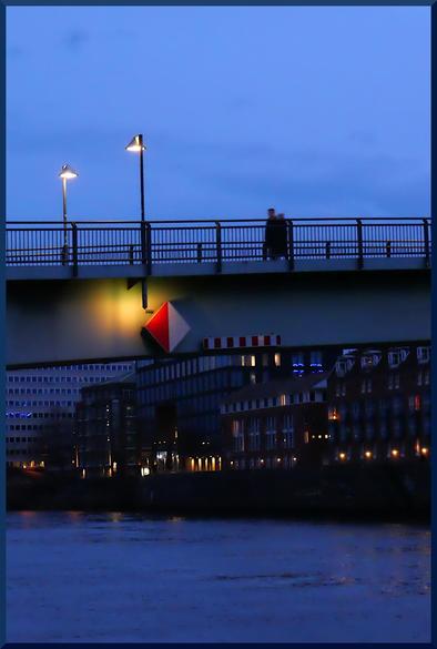 Foto einer Brücke über einem Fluss. Der überwiegende Farbton ist abendliches blau. Zwei Personen gehen über die Brücke. Zwei Laternen spenden ein gelbliches Licht. Ein rot-weißes Schifffahrtszeichen wird von einem gelblichen Licht erleuchtet.
Das Bild wurde in Bremen aufgenommen.