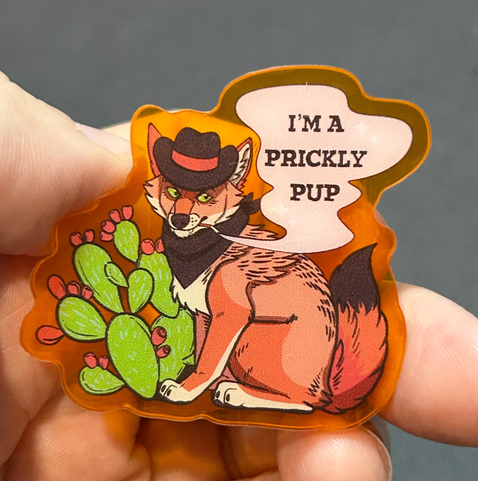 Hand holding an orange acrylic pin of a grumpy coyote pup smoking, wearing a cowboy hat and bandana and sitting beside a prickly pear cacti. It says "I'm a prickly pup"