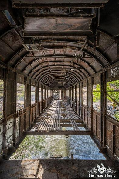 Interior view of an abandoned, rusted train car with missing floor panels, graffiti on the walls, and a river visible below through the gaps.