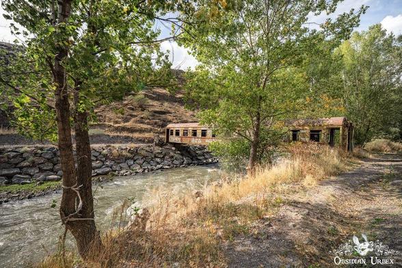 A rusted, abandoned train car sits next to a creek, partially obscured by trees and overgrown vegetation, with a rocky hillside in the background.