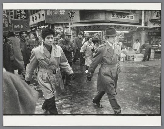 The image is a black and white photograph depicting two men in trench coats, walking briskly through what appears to be an urban street market. The man on the left has dark hair that falls just above his shoulders and wears glasses; he's wearing a light-colored trench coat with visible buttons and belt, as well as trousers tucked into boots. His companion is further ahead of him, sporting a hat and holding papers in one hand while carrying what seems to be a briefcase or bag on the other shoulder.

The street behind them is bustling with activity. Several individuals can be seen engaged in various activities: walking, shopping, some are interacting near market stalls that display goods for sale. The surrounding buildings have signs written in Japanese characters, suggesting this scene takes place in Japan, likely Osaka given the reference to Yakuza-mannen op straat in Osaka.

The pavement is wet and reflective, indicating recent rainfall or a cleaning activity. The overall atmosphere of the photo suggests it could be documenting an event from post-World War II era Japan with its depiction of everyday life amidst economic recovery efforts.

Additional information about this image can be found under Yakuza-mannen op straat in Osaka by Ed van der Elsken, dated 1960.