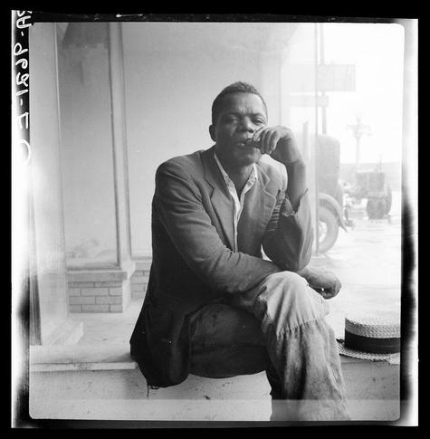 The photograph captures an individual sitting on a low stool or curb outside what appears to be a storefront with large glass windows. The person is dressed in casual attire consisting of a dark, possibly worn-out jacket over light-colored pants and black shoes that show signs of wear at the toes.

They are resting their chin thoughtfully against one hand while the other rests along their body on top of the knee or stool. Their gaze seems directed off to the side rather than towards the camera, suggesting contemplation or introspection.

The setting is an urban environment with visible elements like a brick wall and what looks to be part of a street scene reflected in the window behind them. A straw hat lies unattended on the ground beside the individual, adding a sense of casualness to their demeanor.

This black-and-white photograph carries a historical quality, indicative of its likely mid-20th-century origin. The image is labeled with text at the top left corner that reads "Dorothea Lange" and beneath it in smaller print "1936," suggesting this was photographed by Dorothea Lange during her active years as an American photographer.

The overall atmosphere of the photograph conveys a moment frozen in time, capturing not just the physical presence but also hinting at deeper personal or societal narratives.