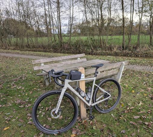 Ein helles Gravelbike steht angelehnt an einem Picknicktisch. Im Hintergrund ein Radweg.