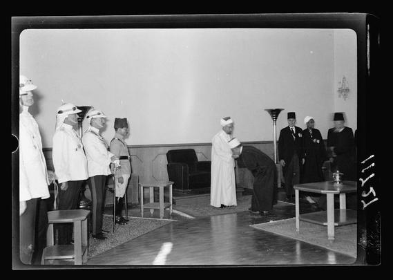 The image depicts a formal gathering of individuals from various backgrounds, dressed in traditional and military attire. The setting appears to be indoors with simple furnishings like chairs, tables, and floor lamps visible. Some people wear white robes and headgear indicative of Islamic religious officials or dignitaries, while others are clad in uniforms suggesting they may represent the Arab Legion. One individual is bowing respectfully towards another who stands prominently among them. The overall atmosphere suggests a significant event with symbolic gestures such as standing attention and respectful greetings signifying honor and respect for those present.