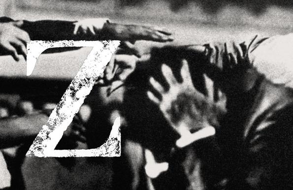 Black and white grainy backdrop featuring a large distressed letter 'Z' on the left side. The blurred image shows a man in profile clutching his head in apparent pain on the right, while hands/arms from a crowd extend toward him from the left side of the frame.