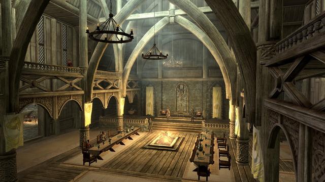 A Skyrim (a videogame) screenshot. A wide-angle, interior view of a large, cavernous hall constructed primarily of dark wood, likely a great hall or similar large communal space. The hall is dominated by a high, arched ceiling supported by numerous wooden pillars and beams, creating a sense of immense scale. Along the center of the hall is a long, rectangular table surrounded by rows of smaller tables on either side, with figures seated at them. In the distance, a raised platform with an ornate chair is visible, with a framed image hanging on the wall behind it. Light sources are distributed throughout the hall, illuminating the wooden structure and casting shadows on the floor and walls.